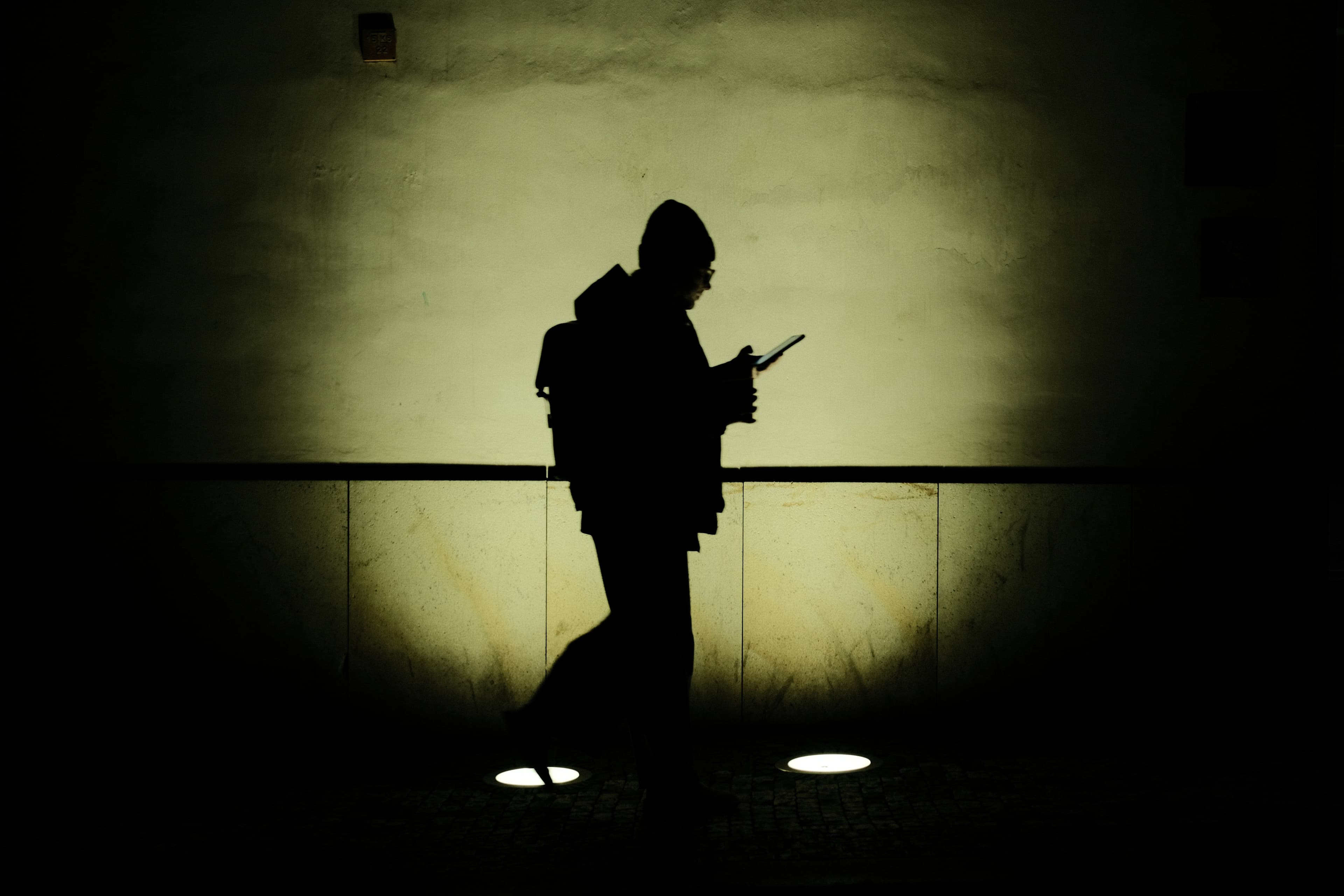 Silhouette einer Person mit Rucksack, die in dunkler Umgebung auf ein Smartphone blickt. Die Person ist von einem hellen Lichtkegel an der Wand hinter ihr umrahmt.